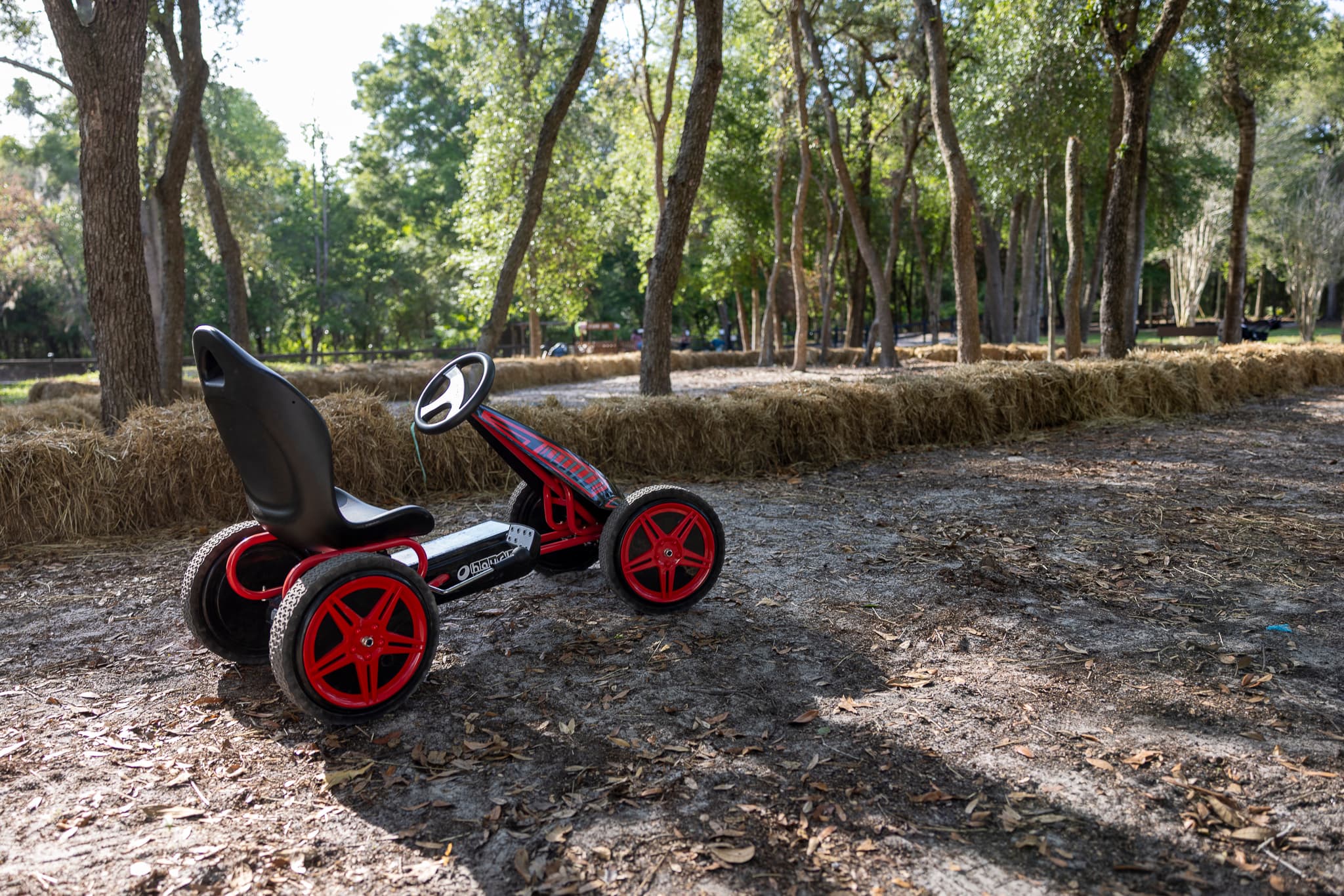 Red and black pedal go-kart on a dirt track lined with hay bales among trees.